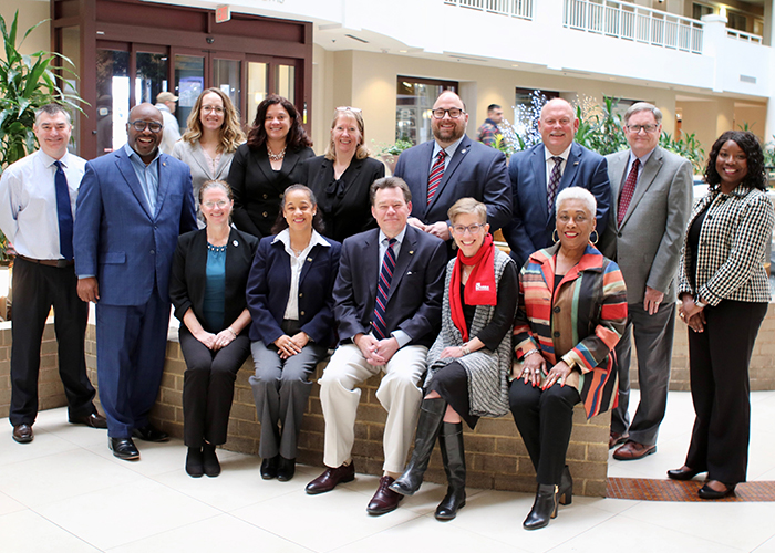 A group of men and women, some sitting on a small brick wall, pose in a hotel atrium.